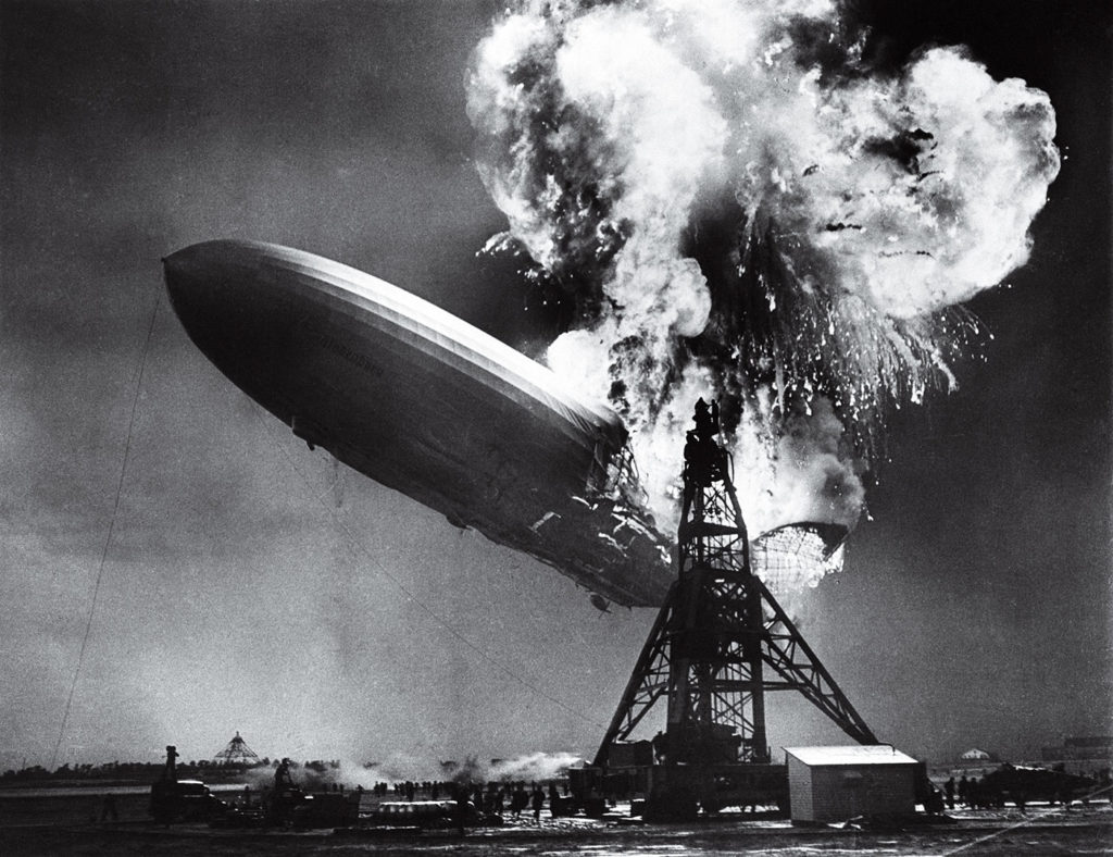 Ein großes Luftschiff schwebt über einer industriellen Struktur und wird von einer massiven Explosion mit viel Rauch getroffen.